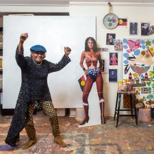 Kaylene Whiskey in her studio, raising her arms in the air in celebration, next to a life size cutout of wonder woman and an artwork in progress