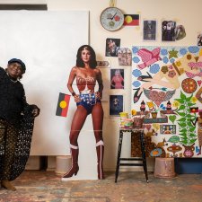Kaylene Whiskey in her studio, raising her arms in the air in celebration, next to a life size cutout of wonder woman and an artwork in progress