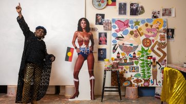 Kaylene Whiskey in her studio, raising her arms in the air in celebration, next to a life size cutout of wonder woman and an artwork in progress