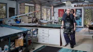 Alison Alder wearing a paint splattered apron, leaning against a bench in her studio