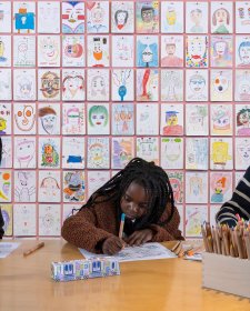 Three people drawing at a wooden table in front of a wall of drawings